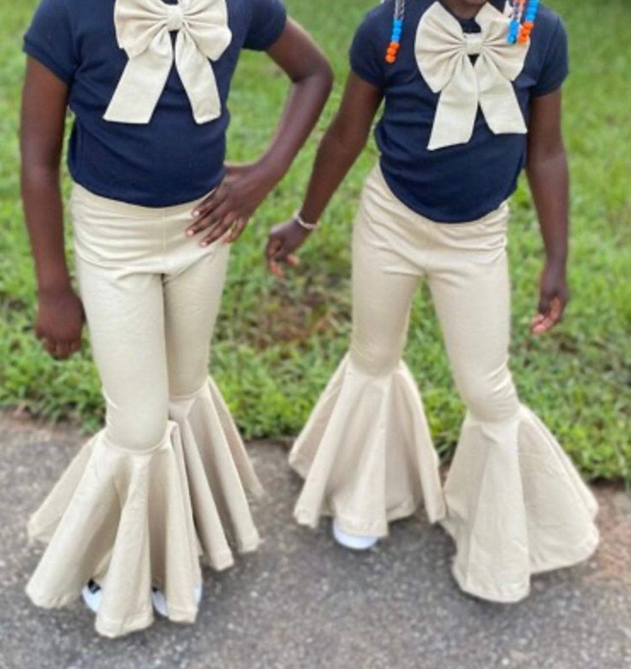 Two children wearing beige school uniform flare pants with navy blue tops and large cream bows outdoors on pavement and grass background